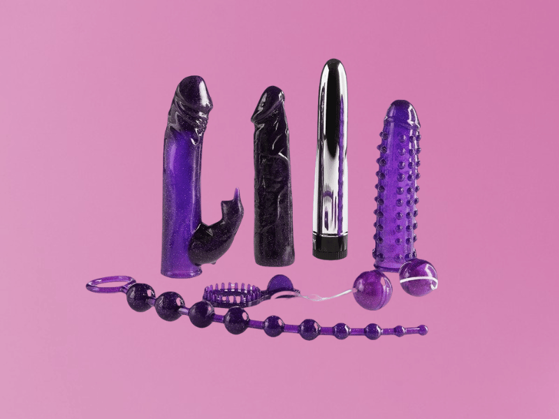 Varietà di sex toys viola e nero su sfondo rosa, inclusi vibratori, dildo con ventosa, palline anali e guaina per pene stimolante